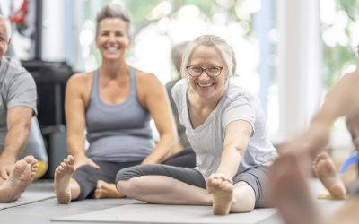 A group of mature adults sit  spread apart on Yoga mats as they stretch out their legs before class.  They are each dressed comfortably in athletic wear and are smiling as they enjoy the fitness class.
